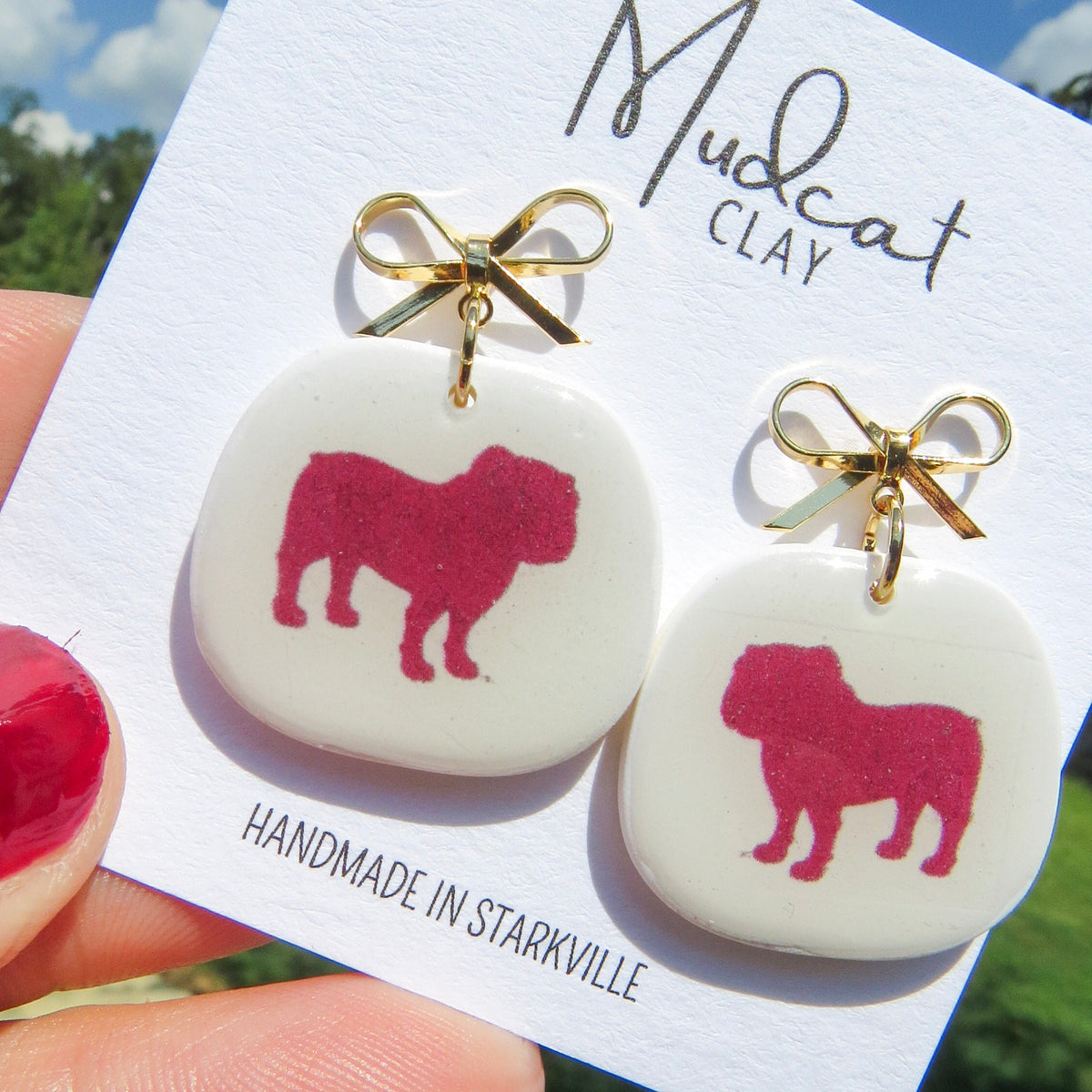 A close-up of a hand holding a white display card with a pair of stud earrings. The earrings have white square puffy charms with a dark pink bulldog silhouette, gold bow tops, and gold posts.